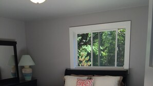 Un dormitorio con cama, ventana con vista a árboles, cómoda con lámpara y alfombra estampada.
