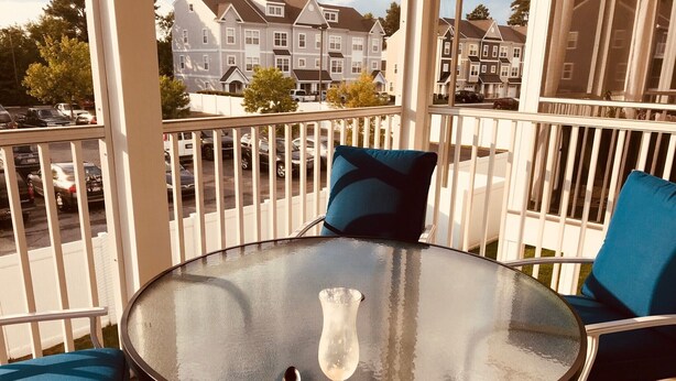 A round glass table with a clear glass vase and a spoon, two blue cushioned chairs, and a white railing on a balcony overlooking a residential street. - Ocean Pointe East - Go to outlets, bike to the beach! (Rehoboth Beach)