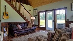 A spacious living room with a wooden staircase, a leather sofa, a guitar, and a large window with a view of greenery.