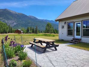 A picnic table in a garden with a mountain view, a wooden cabin, and a building with large windows. - Epic Views - Peaceful, Eco-friendly Home - 30 Minutes to Glacier National Park (Kalispell)