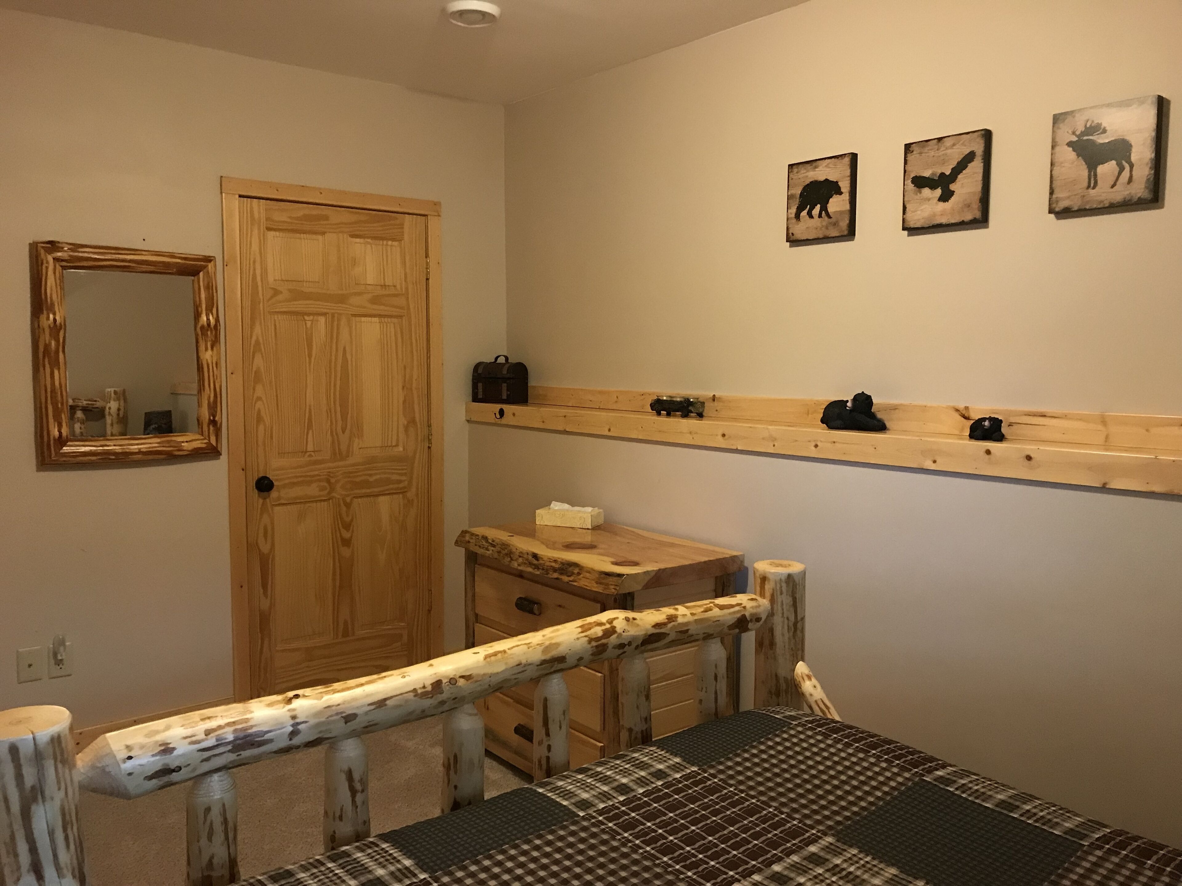 Ein Zimmer mit Holztür, einem Spiegel, einem Bett mit kariertem Überwurf, einem Holz-Kopfteil, einem Regal mit Dekoration und Wandbildern von Tieren.