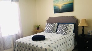 A neatly made bed with patterned bedding, a bedside table with a lamp, a small potted plant, and a painting of a sunset on the wall.