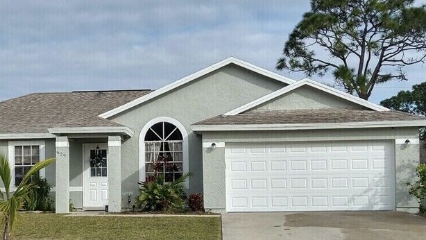A single-story house with a garage, a front door, and a small front yard with a palm tree.