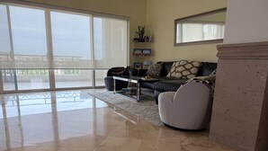 Un salón espacioso con un gran ventanal corredera que da a un balcón, un sofá de cuero negro, un sillón blanco y una mesa de centro.