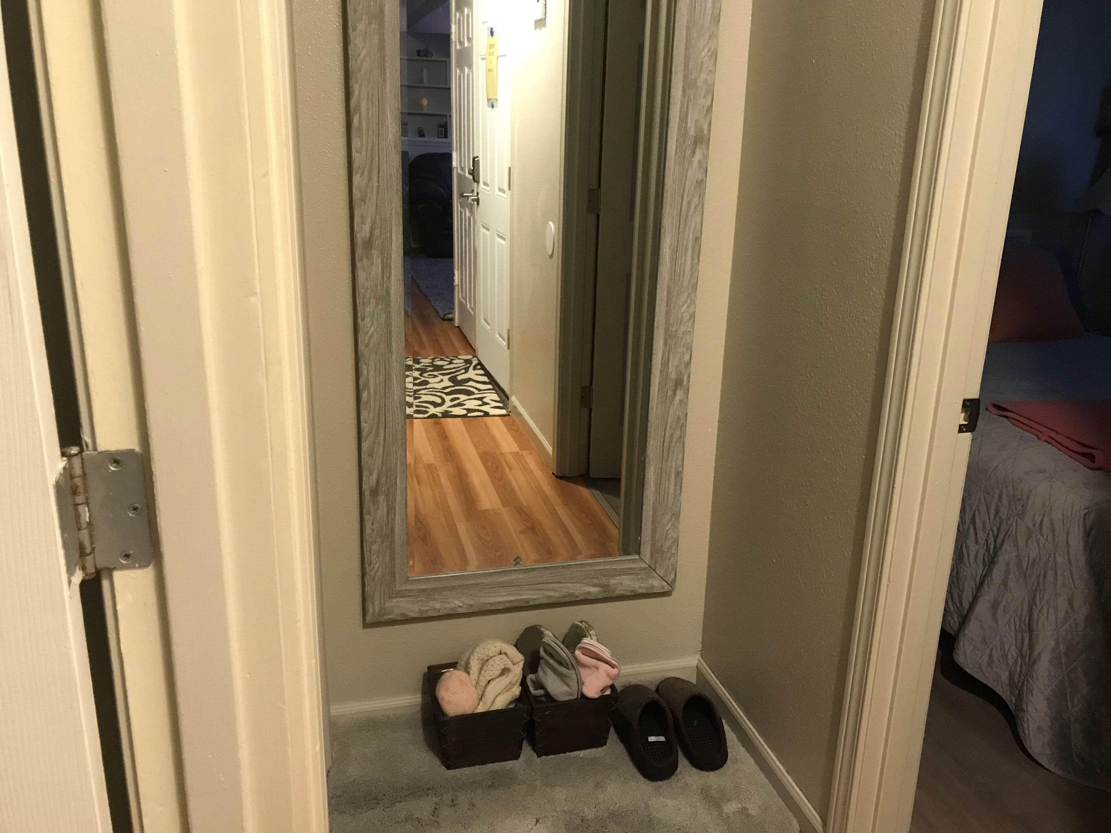 A hallway with a mirror reflecting a room, a door, and a shoe rack with slippers.