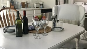 Una mesa de comedor con botellas de vino, copas y un frutero. Hay una silla con cojín, una zona de cocina al fondo y una alfombra en el suelo.