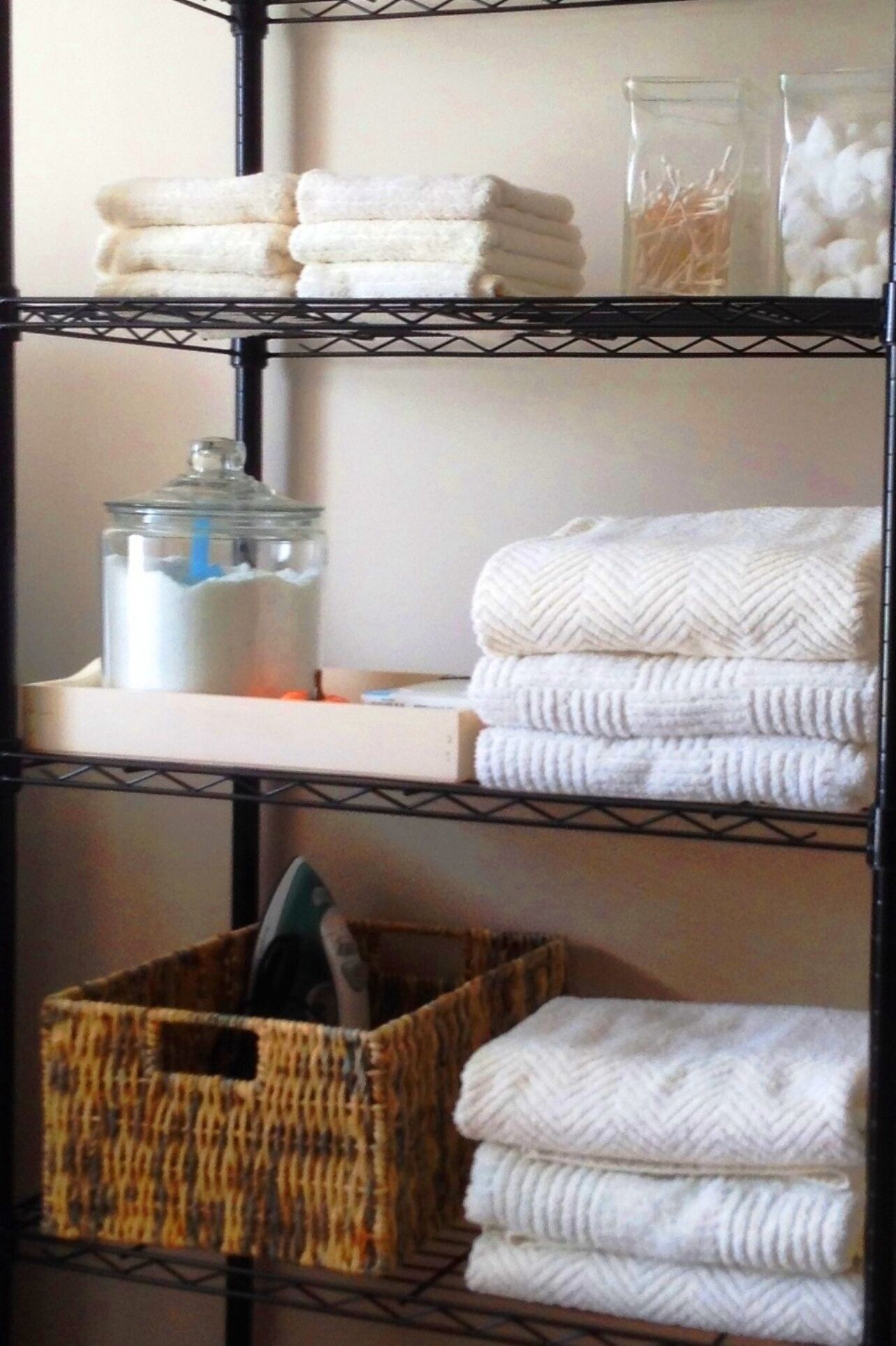 A storage rack with neatly folded white towels, a glass jar with a blue label, and a wicker basket with a black object.