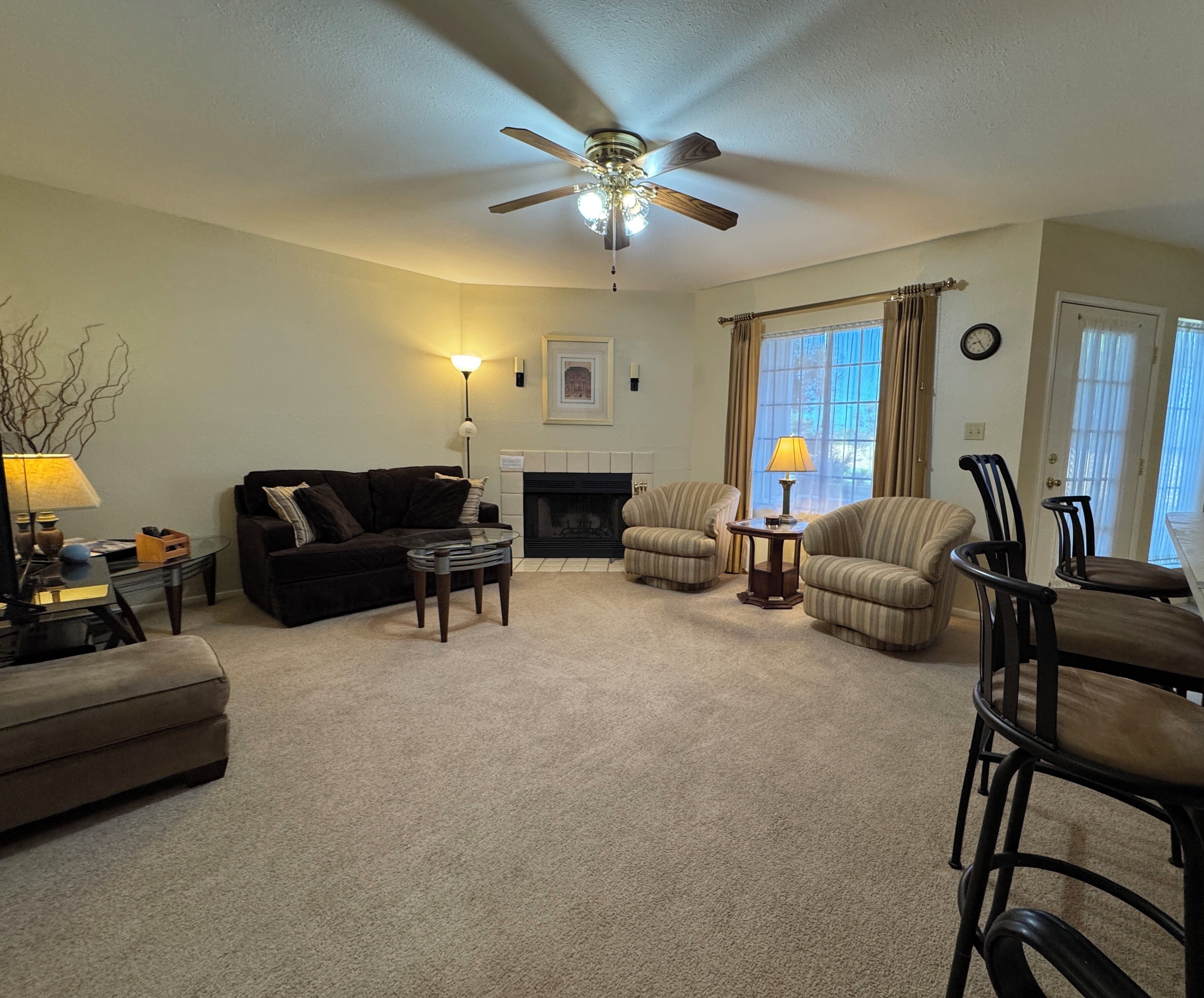 A living room with a ceiling fan, a fireplace, a sofa, a striped armchair, and a dining area with chairs.