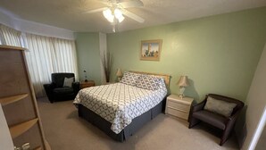 Une chambre à coucher comprenant un lit, deux tables de chevet, une chaise, un ventilateur de plafond et un tableau accroché au mur.