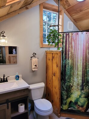 Een badkamer met een houten wastafelmeubel, toilet, wastafel met zeepdispenser, handdoekrek en een raam met uitzicht op bomen.