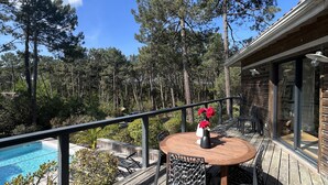 Eine Holzterrasse mit rundem Tisch und Stühlen, mit Blick auf einen Swimmingpool und einen bewaldeten Bereich.