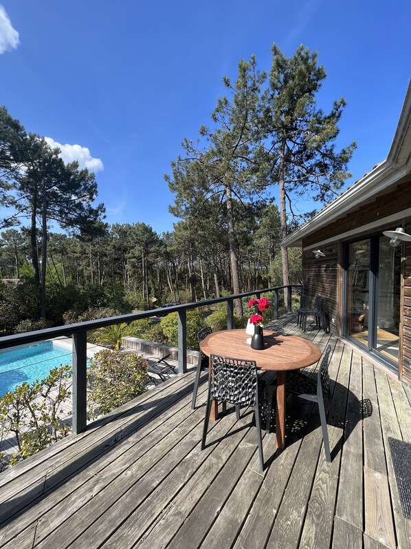 A wooden deck with a round table and chairs, overlooking a swimming pool and a forested area.