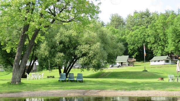 A lakeside park with lounge chairs, a picnic table, and a small building in the background. - Rustic Lakeside Log Cabins (Renfrew)
