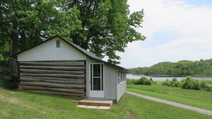A small cabin with a wooden exterior and a white door, situated next to a grassy area with a path leading to a lake. - Rustic Lakeside Log Cabins (Renfrew)