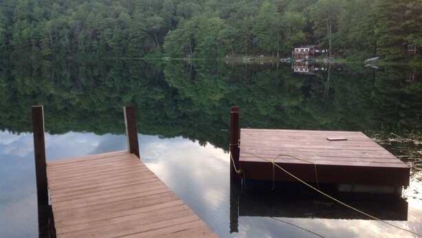 A wooden dock extending into a calm lake, surrounded by dense forest and a small building in the background. - Ultimate relaxing vacation  (Orwell)