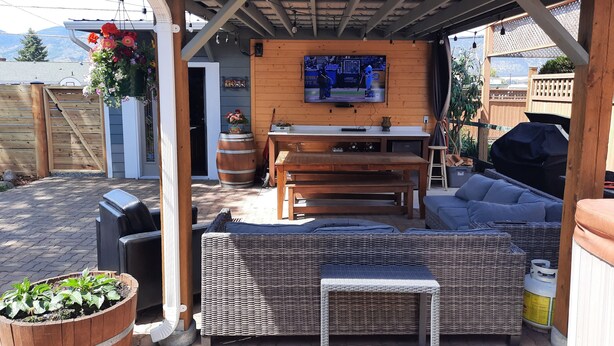 An outdoor patio with a wooden table, wicker sofa, and a TV mounted on a wooden wall.