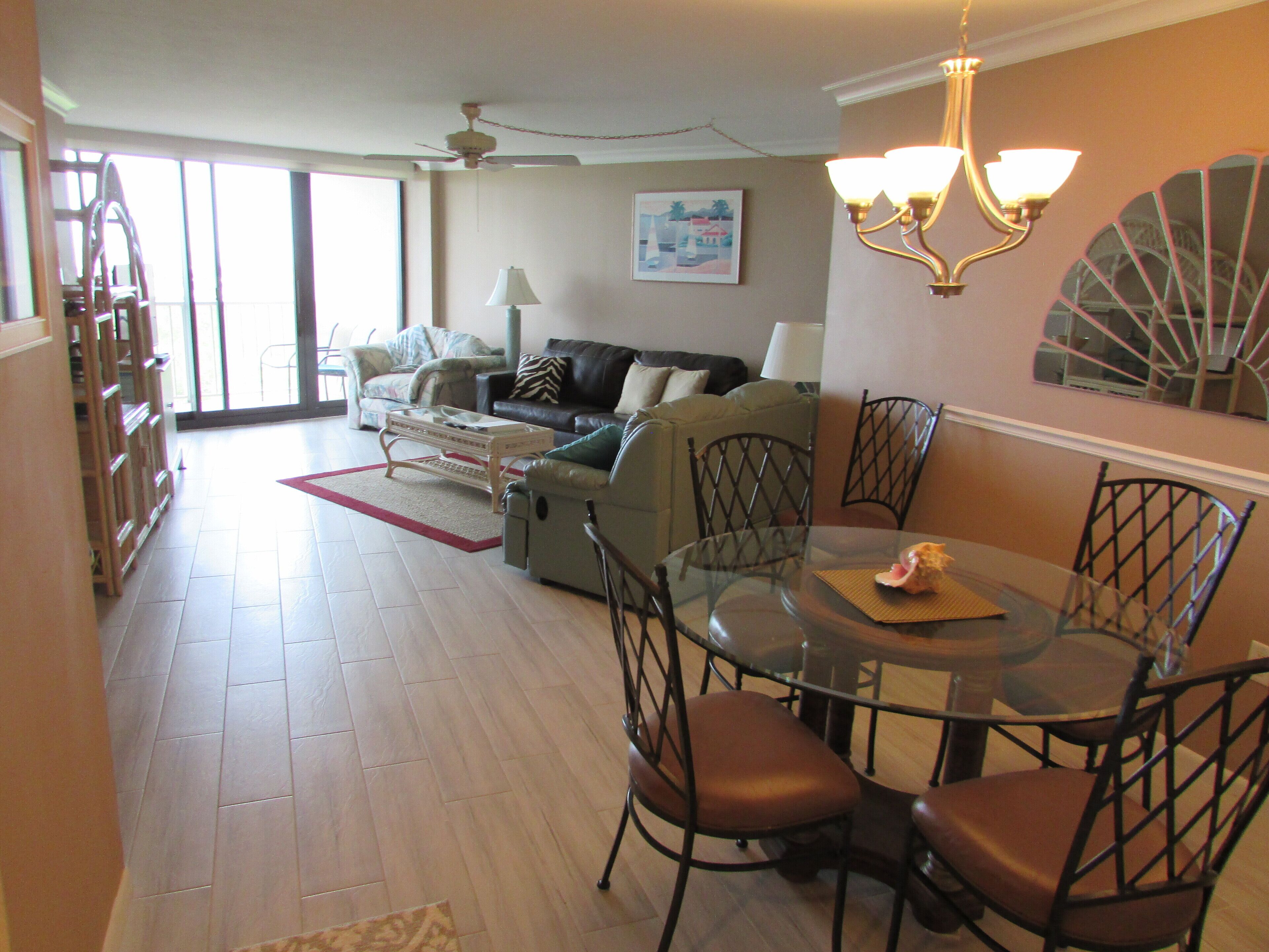A spacious living area with a dining table, chairs, and a sofa. There is a ceiling fan, a lamp, and a painting on the wall.