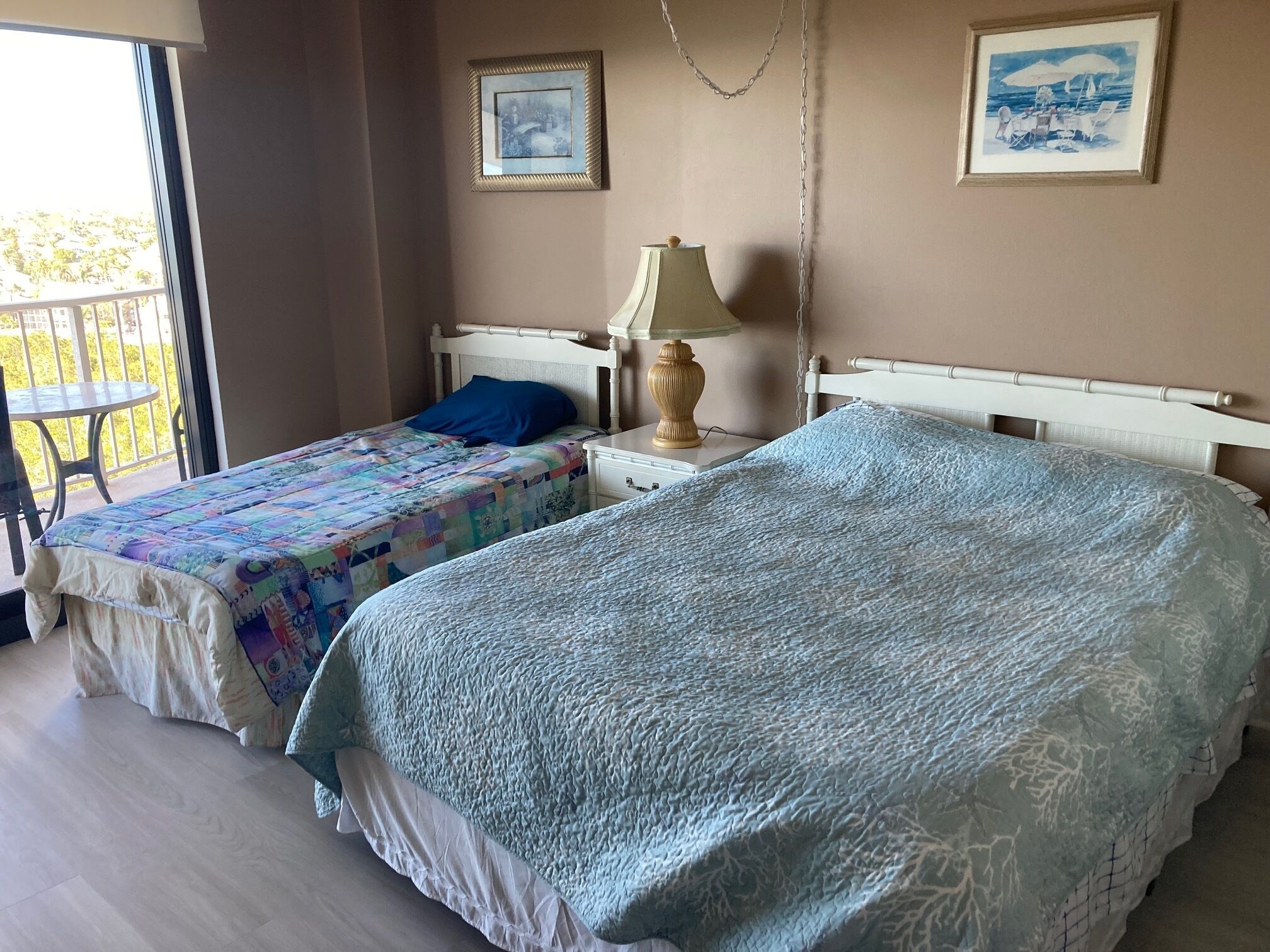 Un dormitorio con dos camas, una mesita de noche con una lámpara y un balcón visible a través de una puerta abierta.