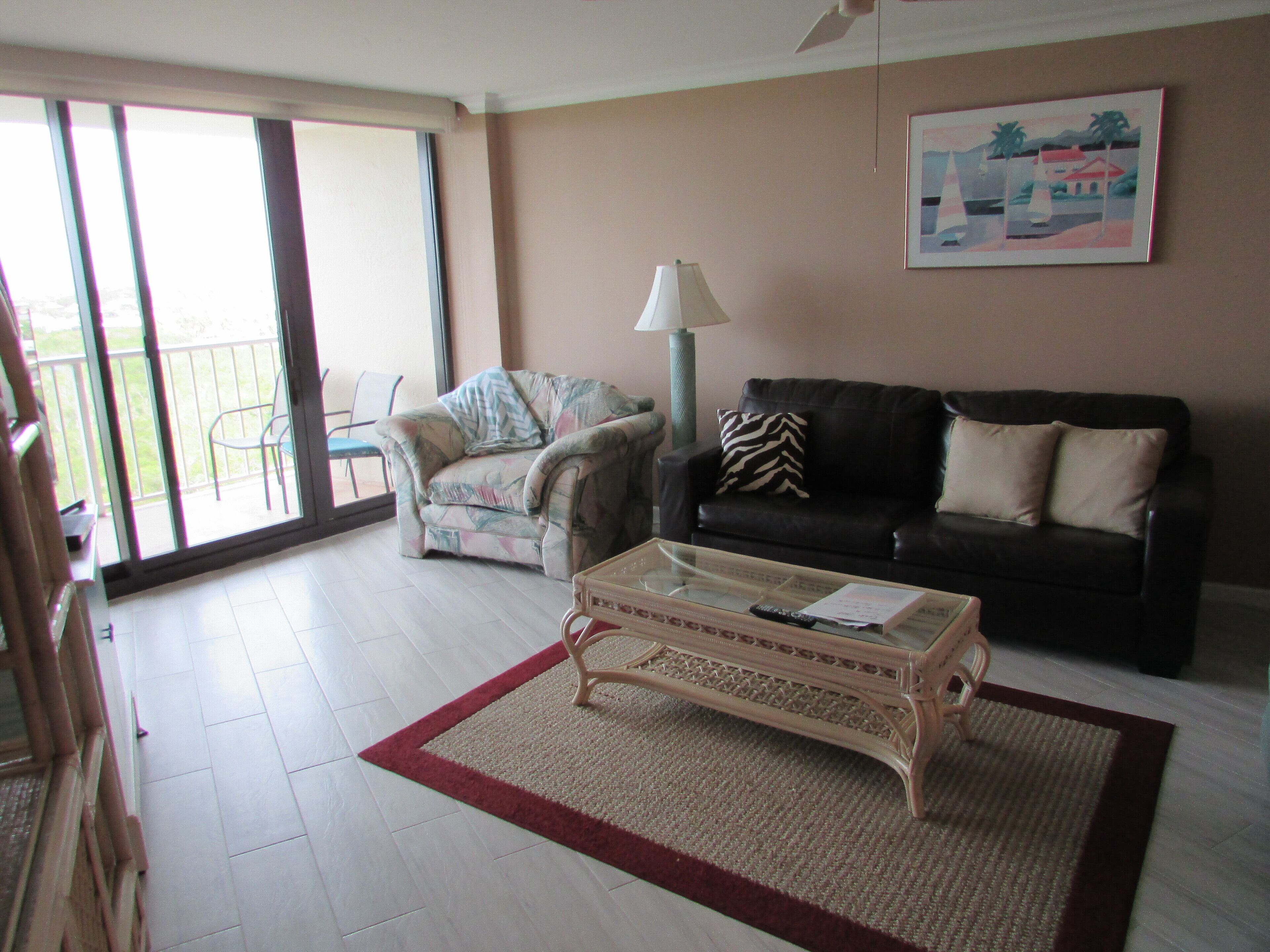 Salón con sofá, sillón y mesa de centro. Cuenta con un balcón con sillas y una lámpara.