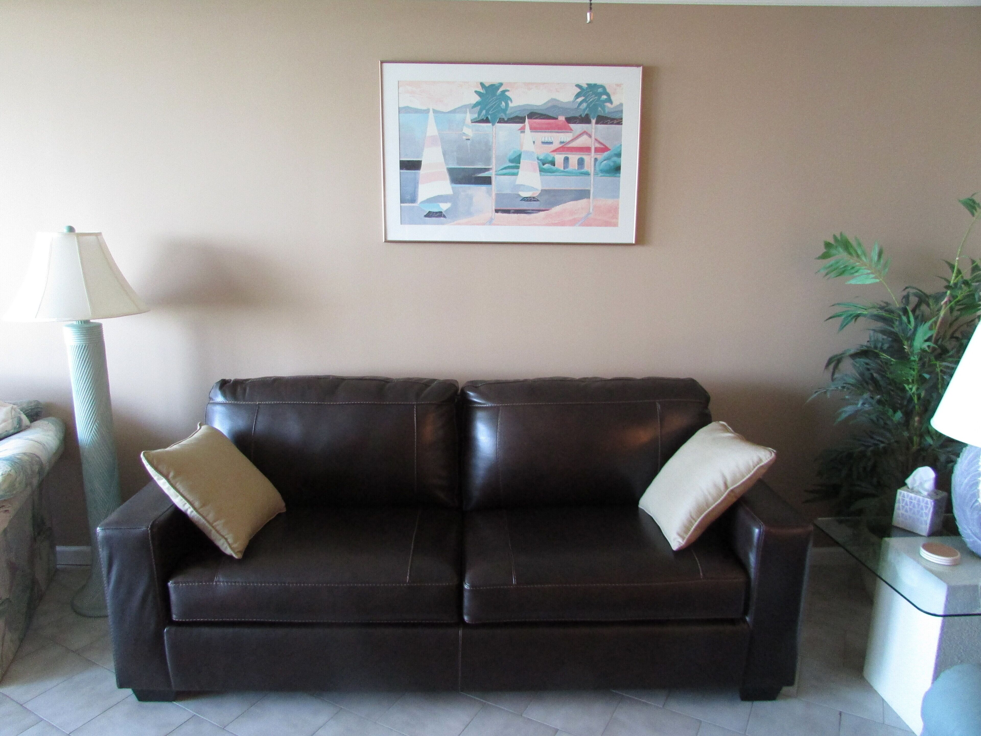 A dark brown leather sofa with two beige cushions, a framed abstract painting above it, a tall floor lamp, and a potted plant to the right.