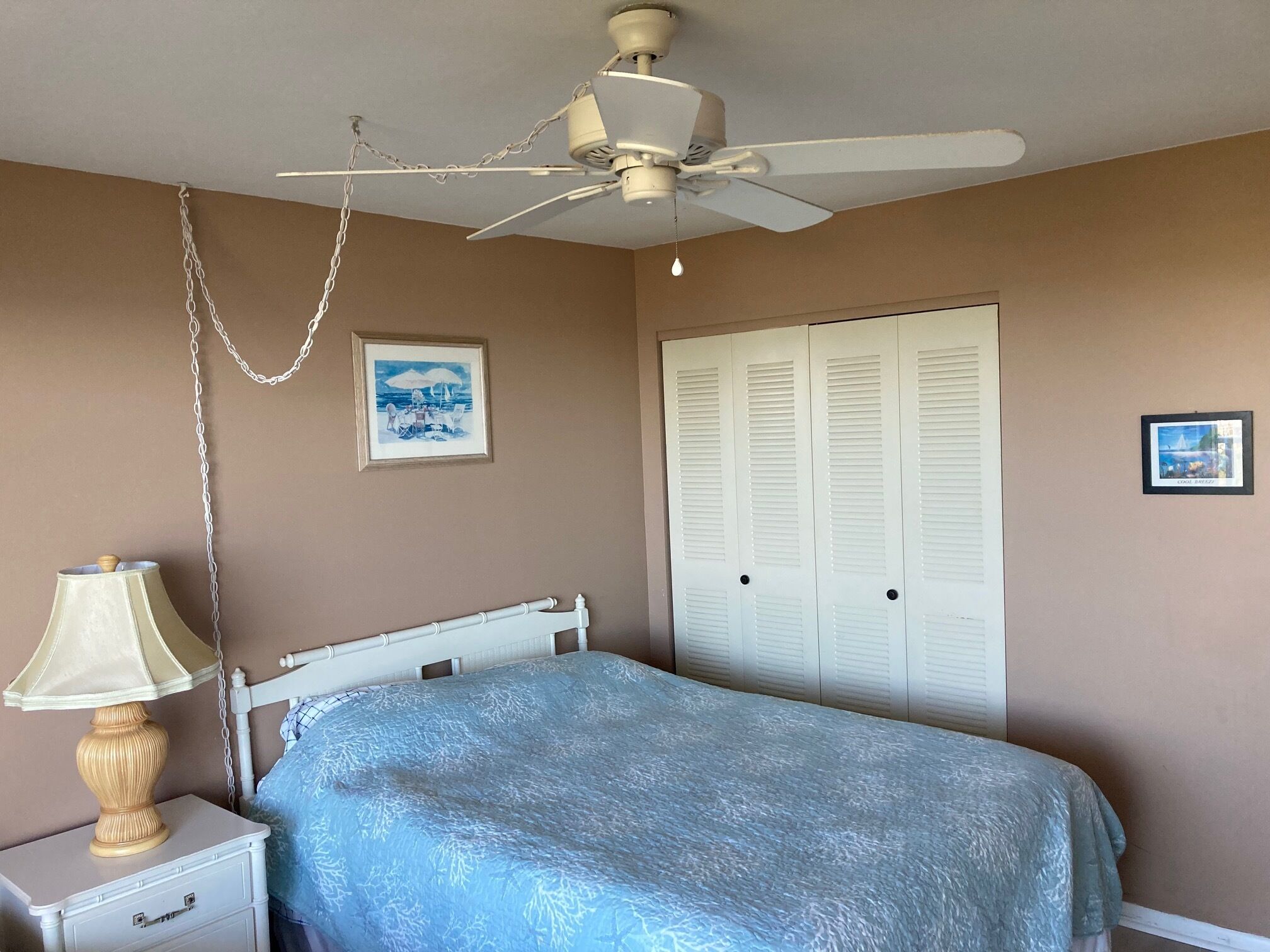 A bedroom with a bed, a nightstand with a lamp, a ceiling fan, and two framed pictures on the wall.