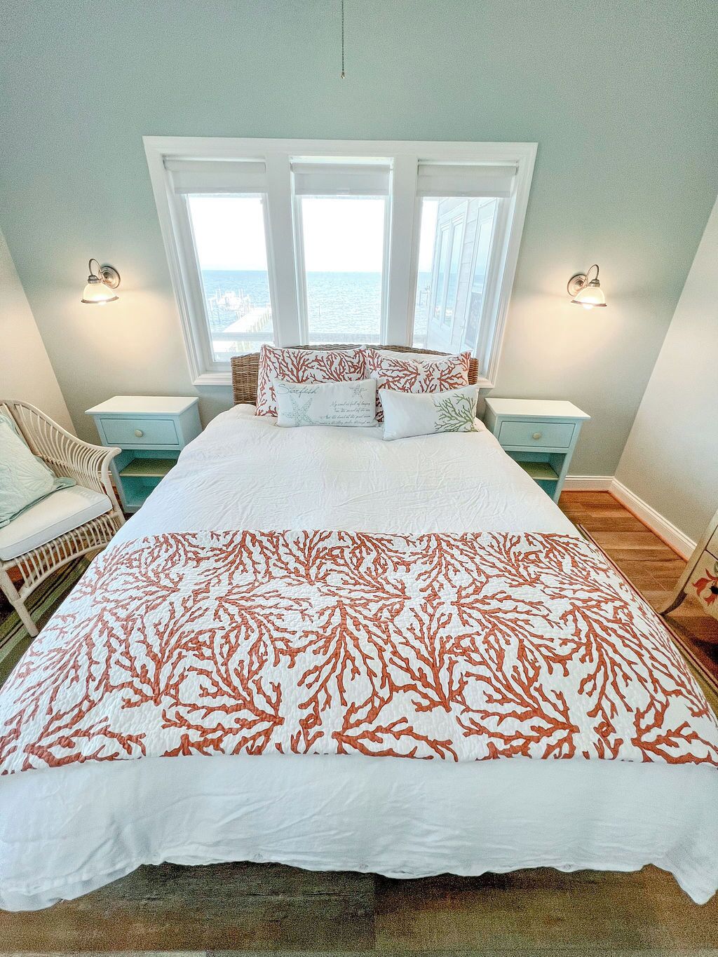A bedroom with a large bed, two nightstands, a wicker chair, and a view of the ocean through three windows.