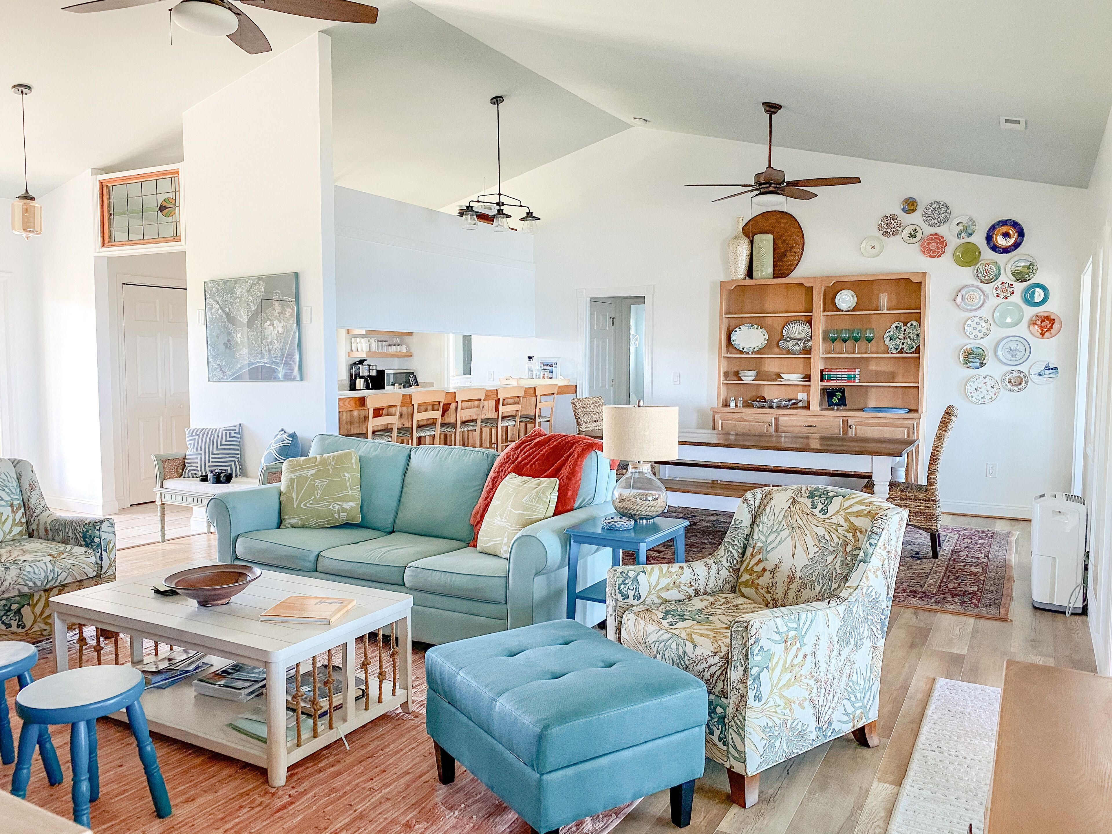 A spacious living room with a light blue sofa, a coffee table, and a patterned armchair. A dining area is visible in the background with wooden furniture and a kitchen area.