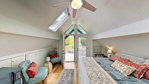 A cozy attic bedroom with a sloped ceiling, a bed with patterned bedding, a seating area with a sofa and chairs, and a view through a door to the outside. - Haleakala Sunrise Home, Kula, Maui, Hawaii (Kula)