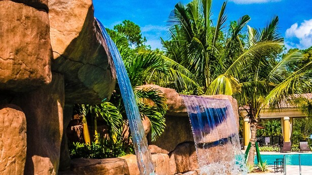 A pool with a waterfall feature surrounded by palm trees and a clear blue sky. - Escape the coldest and snowiest winter in years by "Springing" into Treviso Bay! (Naples)