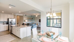 A modern kitchen with white cabinets, a stainless steel refrigerator, and a dishwasher. There is a dining area with a glass table and chairs, and a large window with a view of the city. - Escape the coldest and snowiest winter in years by "Springing" into Treviso Bay! (Naples)