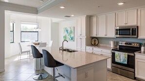A modern kitchen with white cabinetry, a central island with bar stools, stainless steel appliances, and a large window providing natural light. - Escape the coldest and snowiest winter in years by "Springing" into Treviso Bay! (Naples)