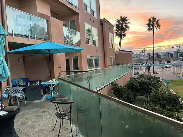 A balcony with a table and chairs, a blue umbrella, and a view of palm trees and a sunset.