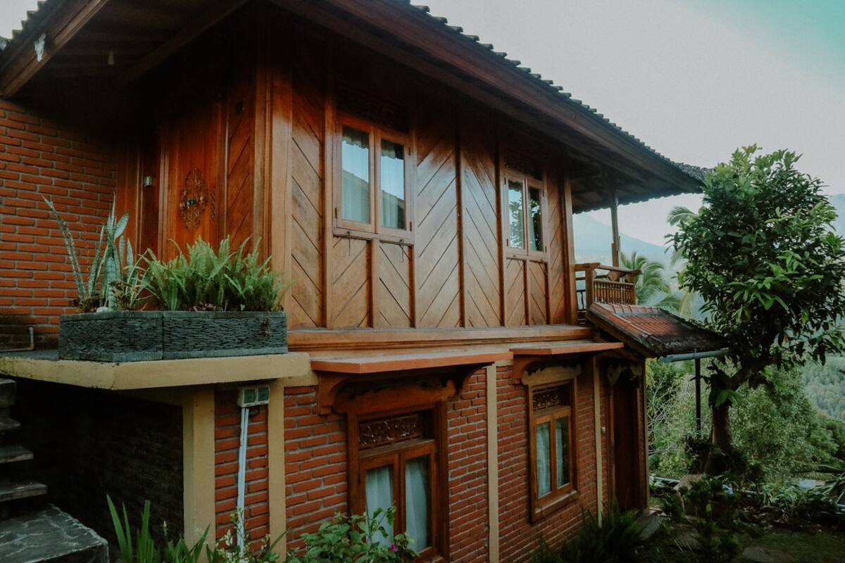 Een houten huis met een balkon, omringd door weelderige begroeiing en een bergachtige achtergrond.