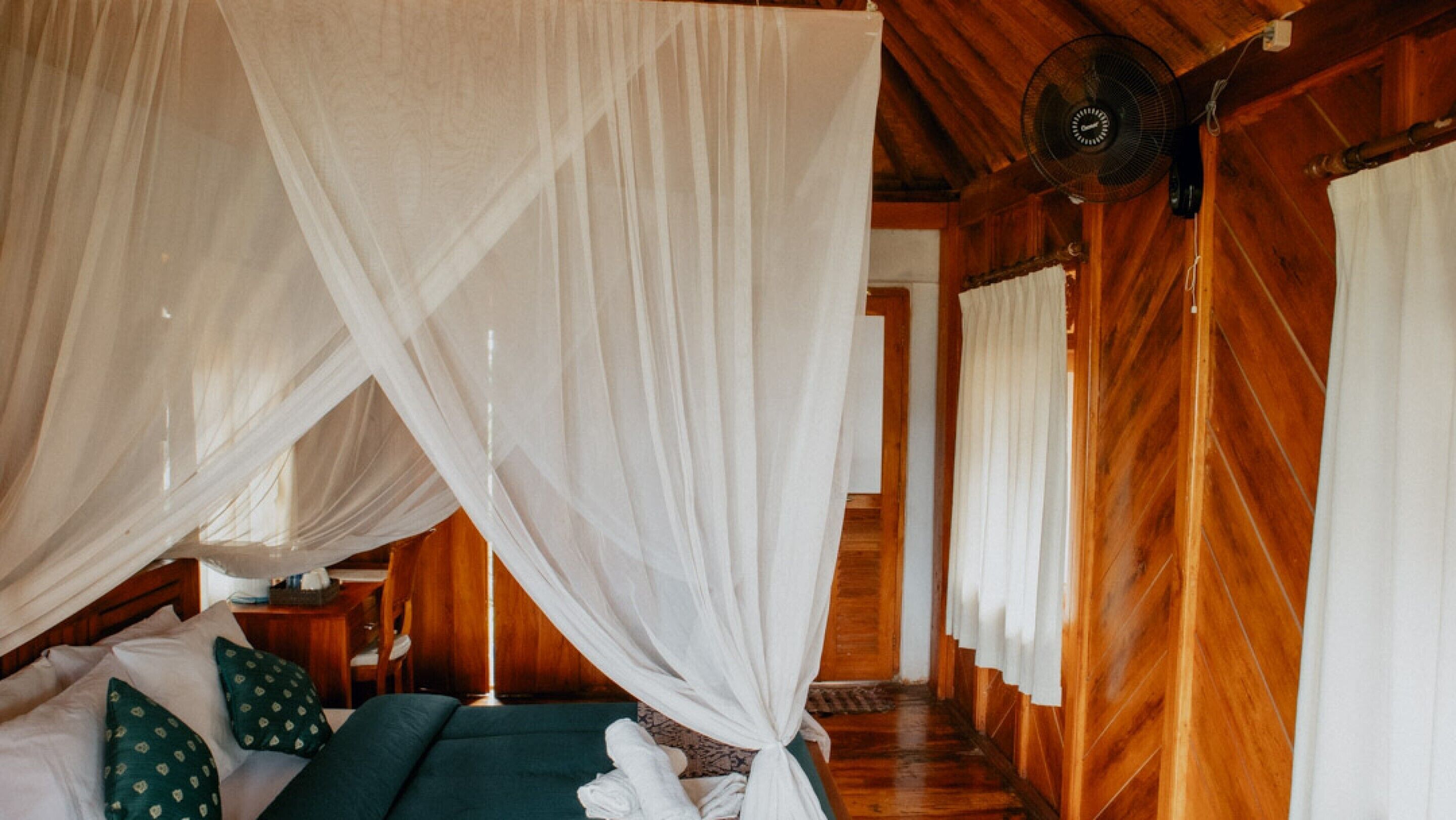 A wooden room with a bed under a canopy, a fan, and a wooden floor.
