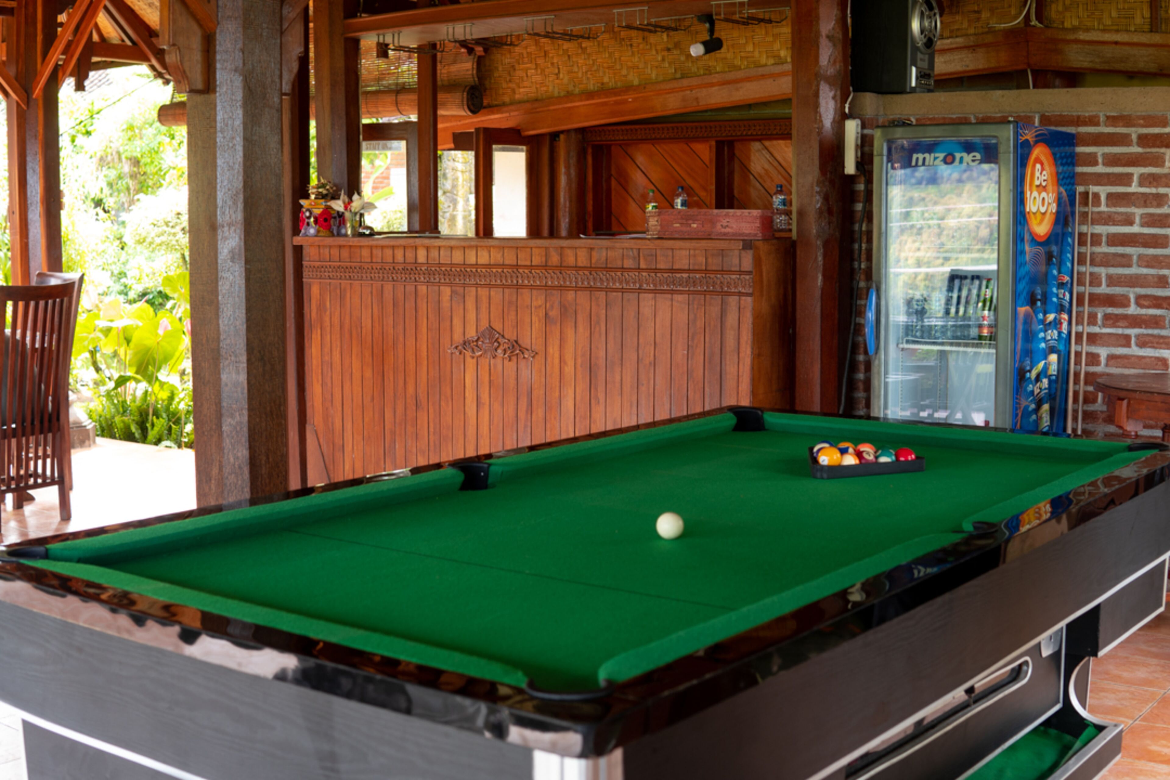 A pool table with balls, a beverage dispenser, and a bar area with a brick wall and wooden decor.