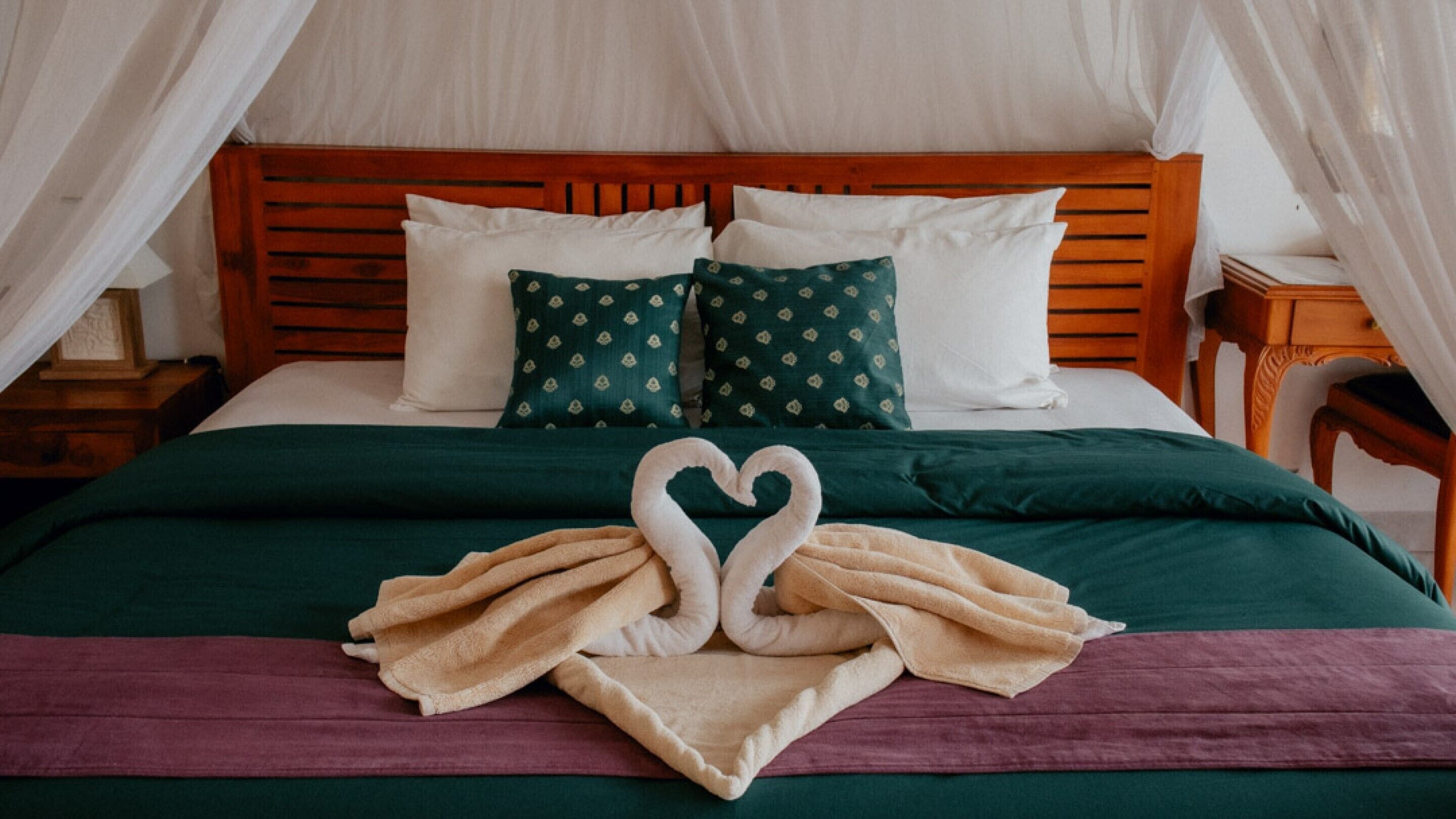 A neatly made bed with a heart-shaped towel arrangement, white and green patterned pillows, and a wooden headboard.