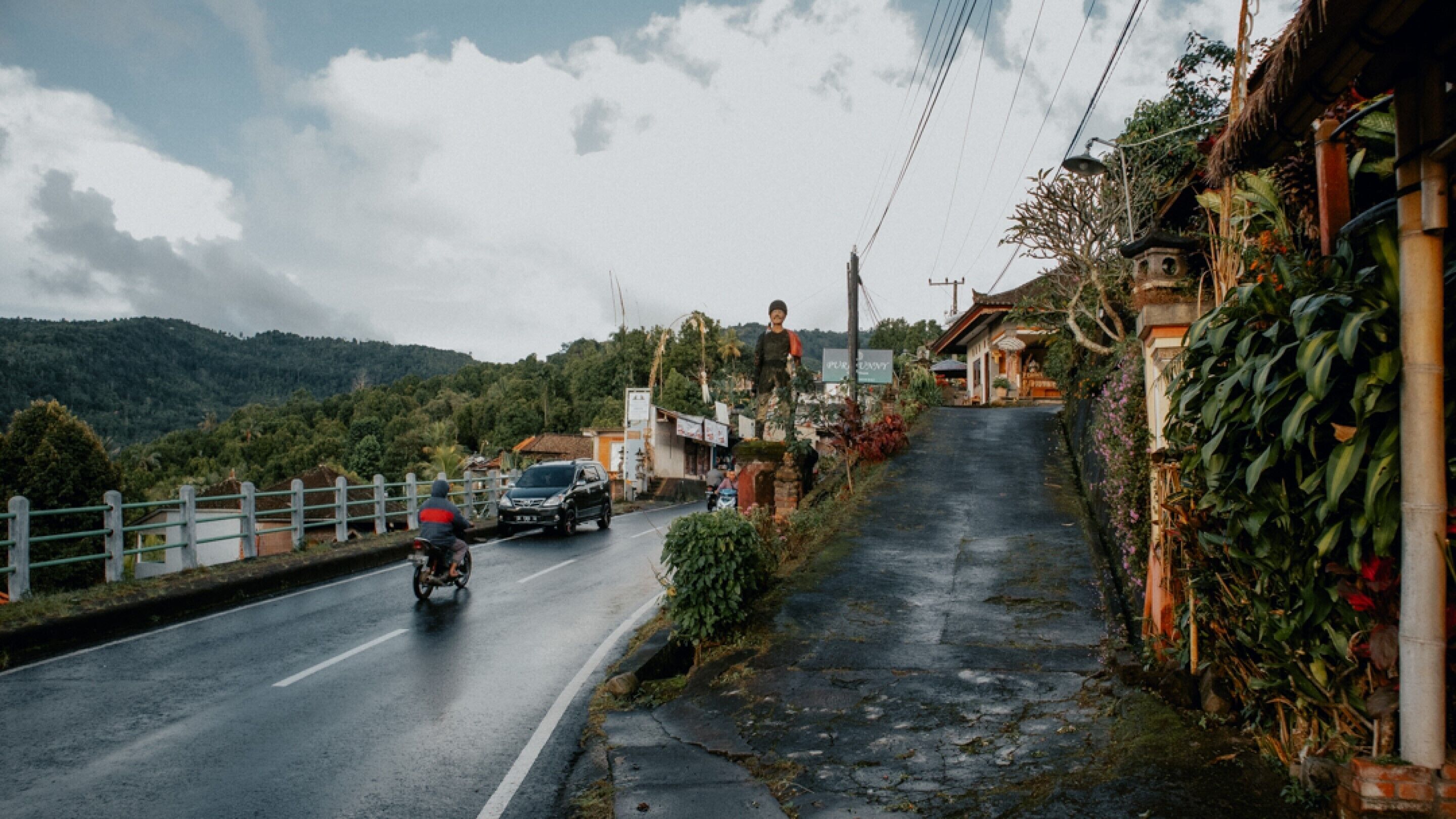 A road with a motorcycle and a car. There are houses and vegetation on the sides.