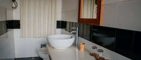Un baño moderno con un lavamanos blanco de diseño, un inodoro, un espejo y una pequeña bandeja de madera con artículos de aseo.