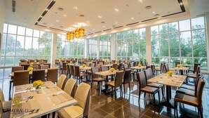 A spacious dining area with multiple tables and chairs, large windows offering a view of greenery, and modern pendant lighting. - Amerald Resort Hotel (Pengerang)