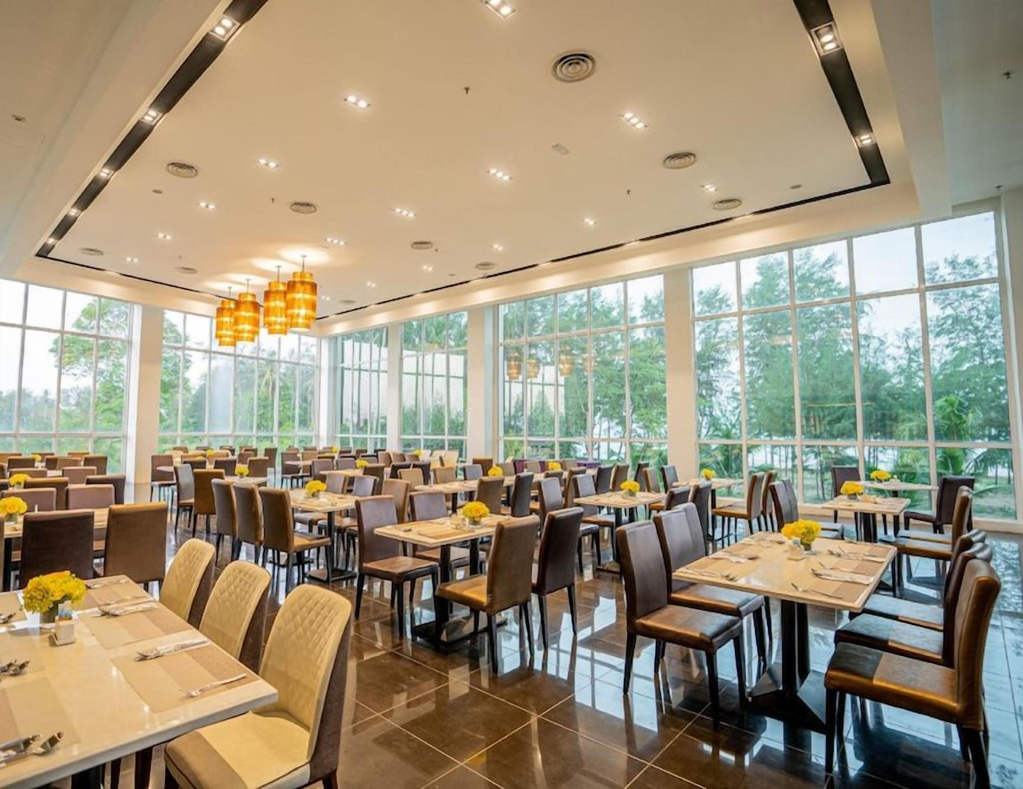 A spacious dining area with multiple tables and chairs, large windows offering a view of greenery, and modern pendant lighting.