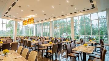 A spacious dining area with multiple tables and chairs, large windows offering a view of greenery, and modern pendant lighting.