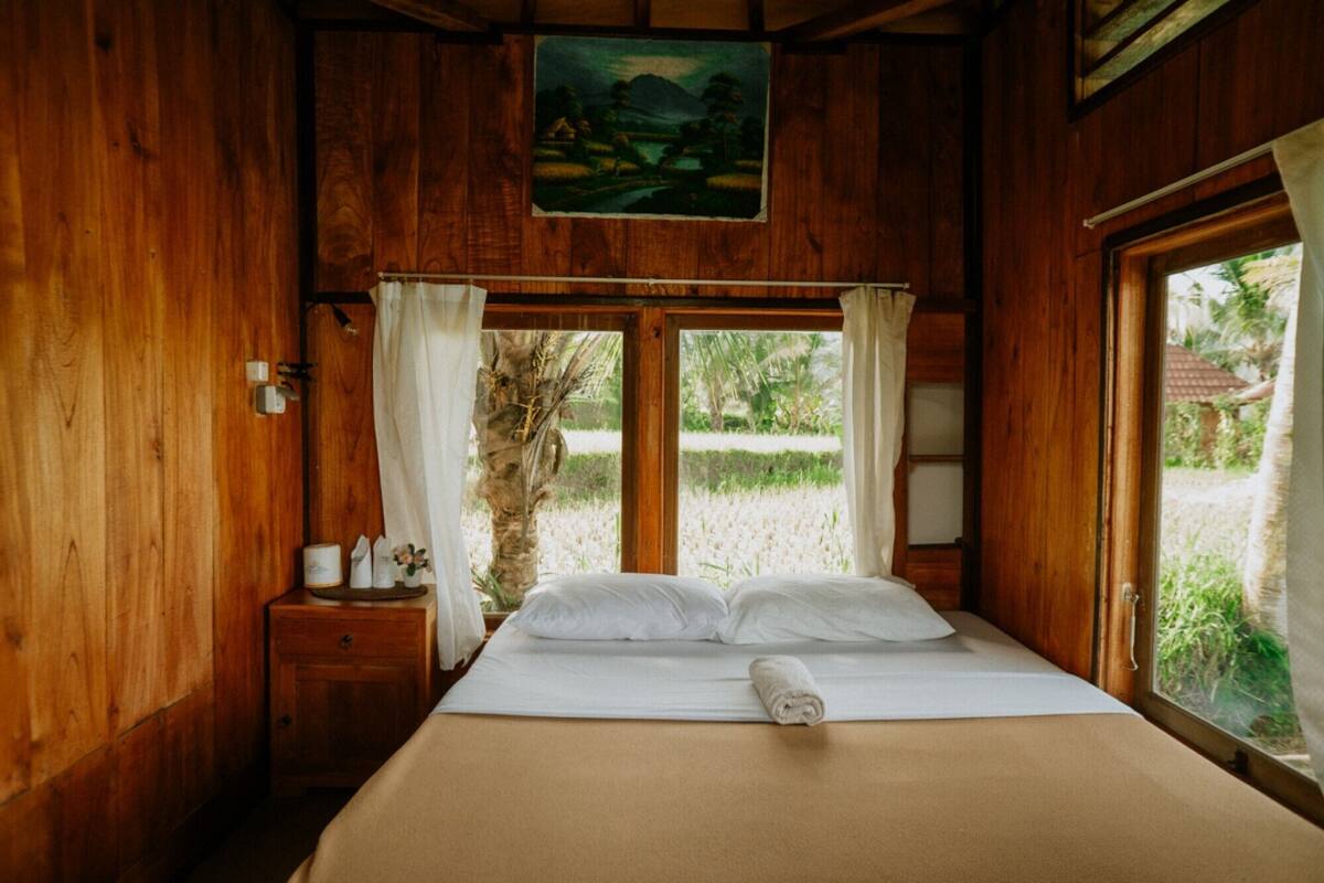 A wooden cabin bedroom with a bed, a nightstand, a painting, and a window with curtains.