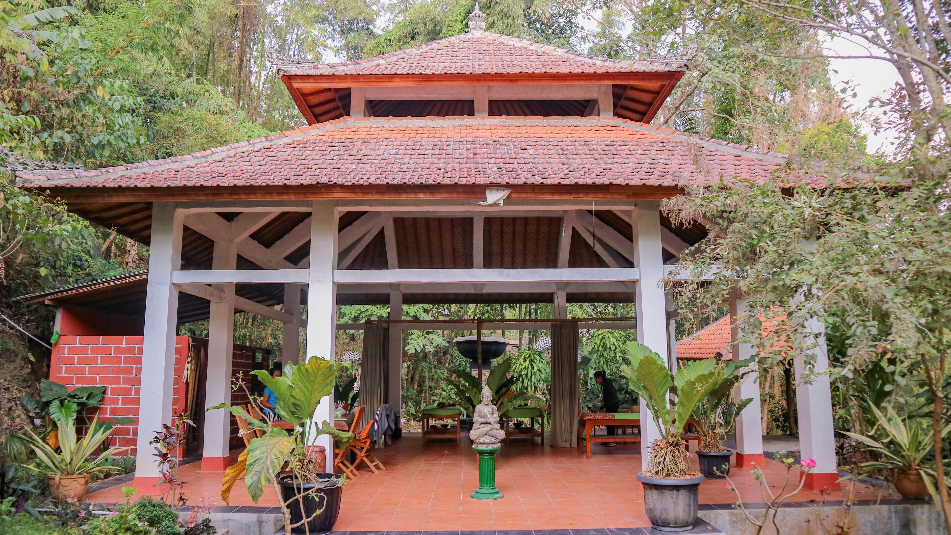 A covered pavilion with a tiled roof, surrounded by potted plants and a Buddha statue.