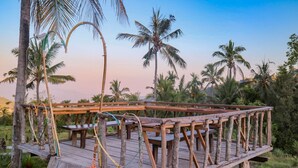 A wooden deck with tables and chairs, surrounded by palm trees and greenery. - Puri Sunny Camping (Munduk)