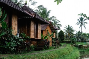 A rustic wooden house with a thatched roof, surrounded by lush greenery and palm trees. - Puri Sunny Camping (Munduk)