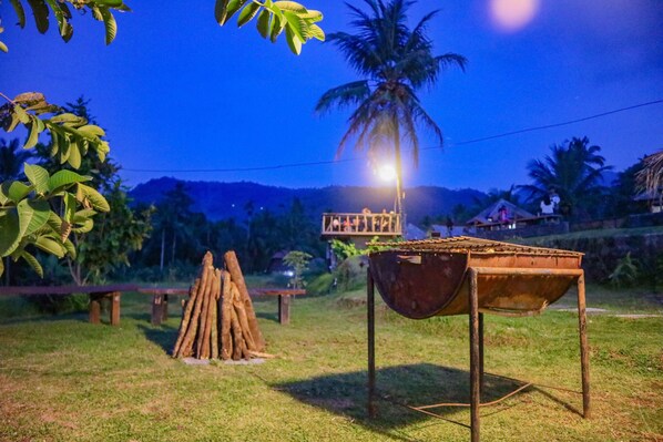 Un décor extérieur rustique, avec une table en bois et un pot suspendu, entouré de verdure et d’un palmier, sous un ciel nocturne.