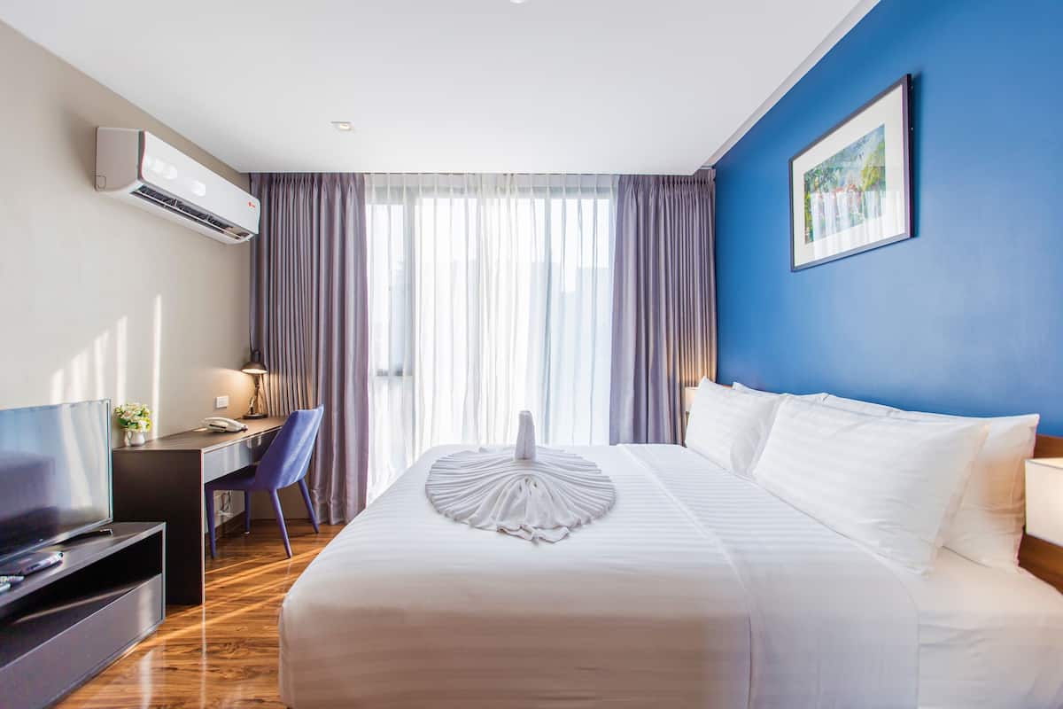 A hotel room with a bed, a desk, a chair, a TV, and an air conditioning unit.