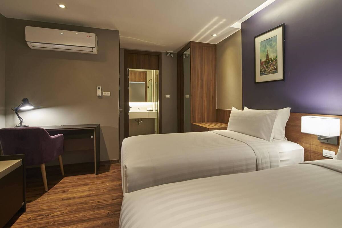 A hotel room with two beds, a desk, a chair, an air conditioner, a mirror, and a painting on the wall.