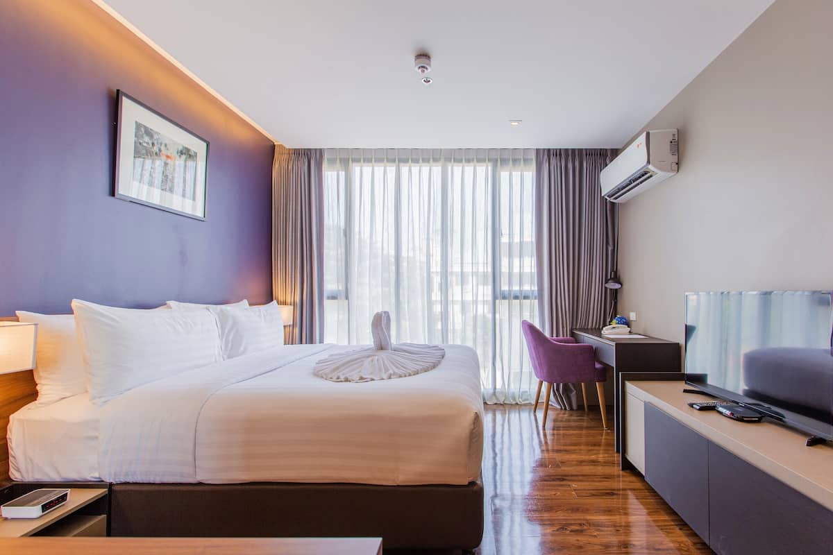 A hotel room with a bed, a desk, a chair, a TV, and an air conditioning unit.