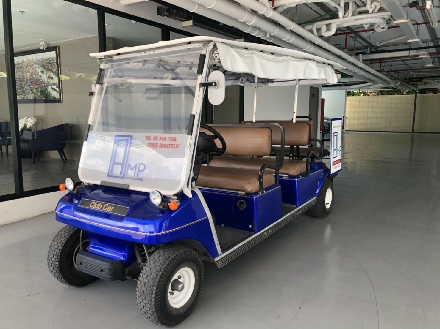 Un carro de golf azul con toldo blanco, identificado como 'Club Car', estacionado en un espacio interior con pared de vidrio y un cuadro en el fondo.